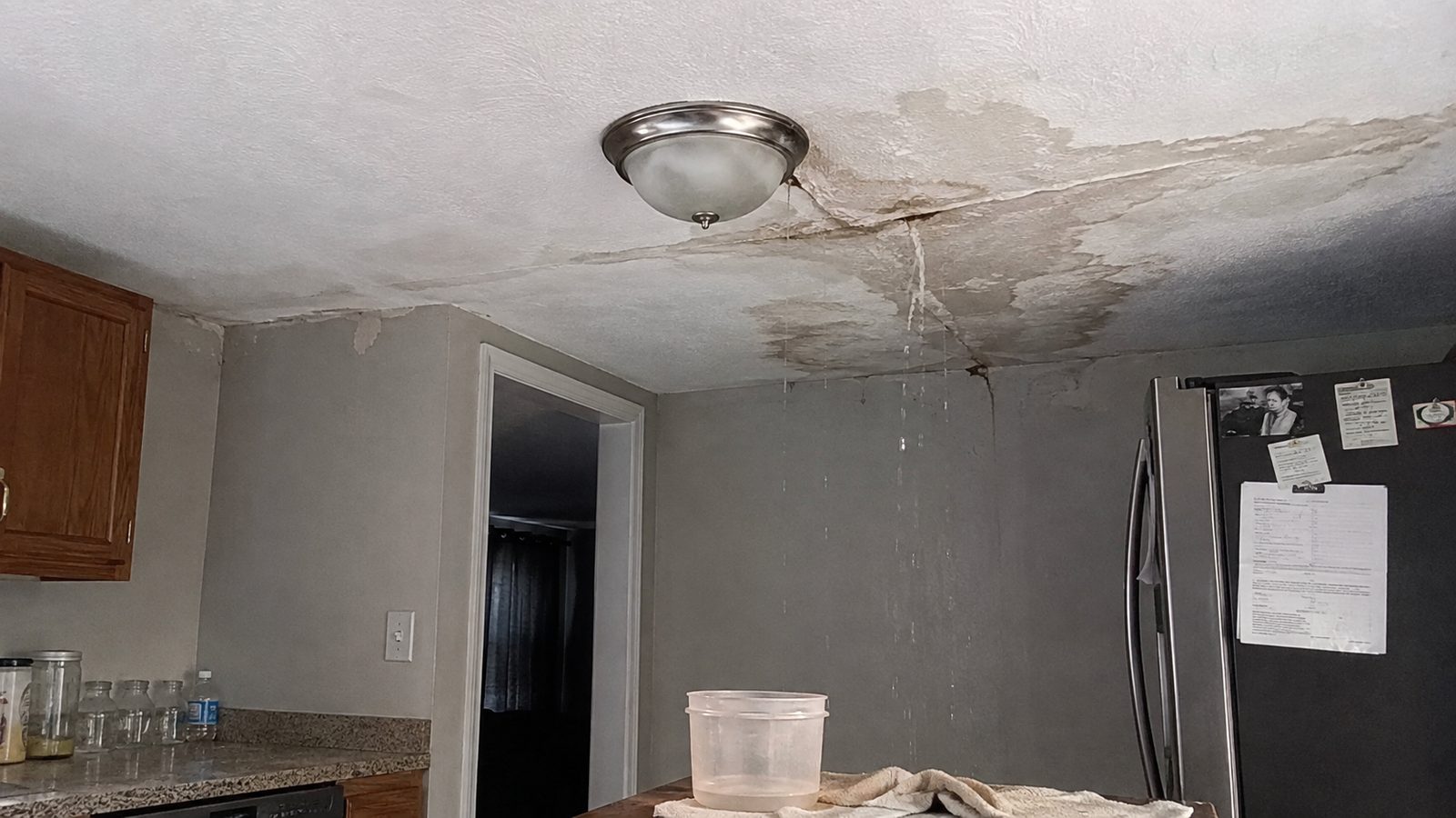 Water-stained kitchen ceiling with an active drip from a roof leak, caught in a container on the counter below.